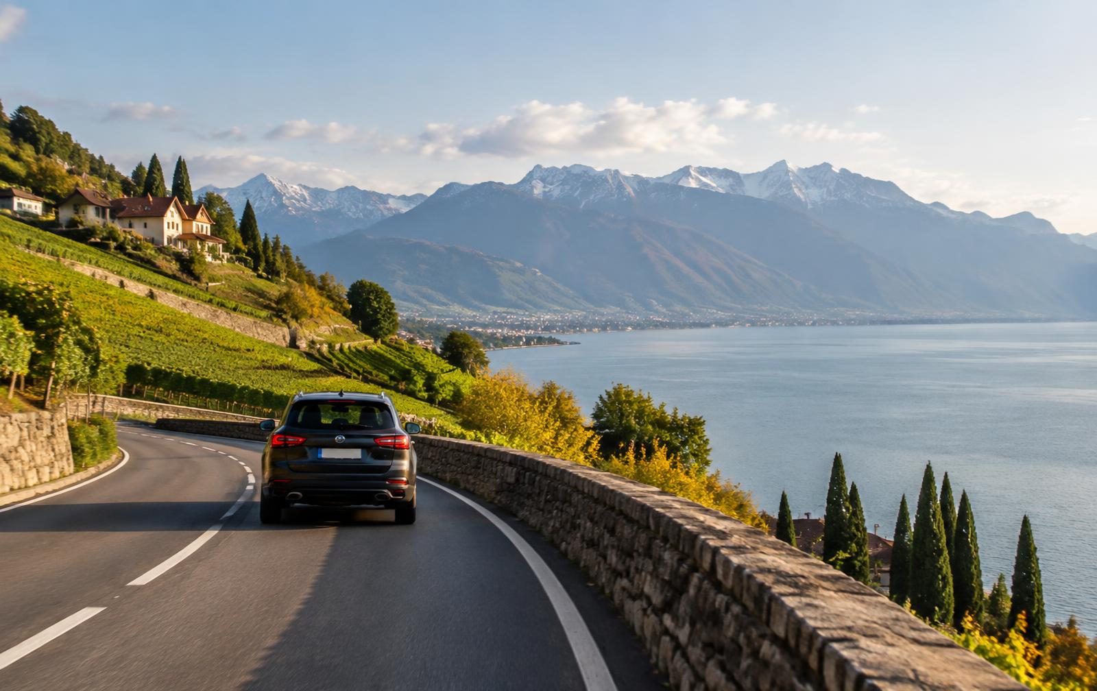 Auto fährt auf einer Uferstrasse am Genfersee vorbei an Weinbergen, im Hintergrund verschneite Berge